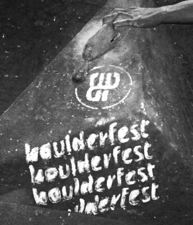 A climber’s chalked hand grips a hold on a climbing wall marked with repeated “boulderfest” text and a circular climbing gym logo. The image is in black and white.