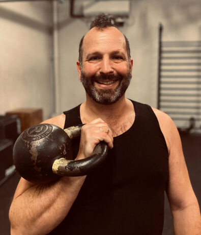 A smiling man with a beard and mustache, wearing a black tank top, holds a worn kettlebell on his shoulder in a gym setting with exercise equipment visible in the background.