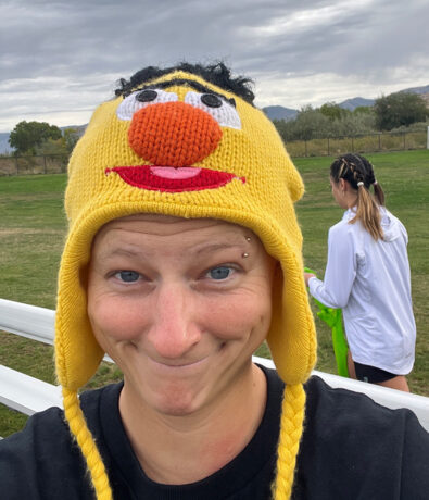 A person smiling and wearing a yellow knit hat with a cartoon face, resembling Bert from Sesame Street, stands outdoors. In the background, another person faces away near a grassy field and cloudy sky.