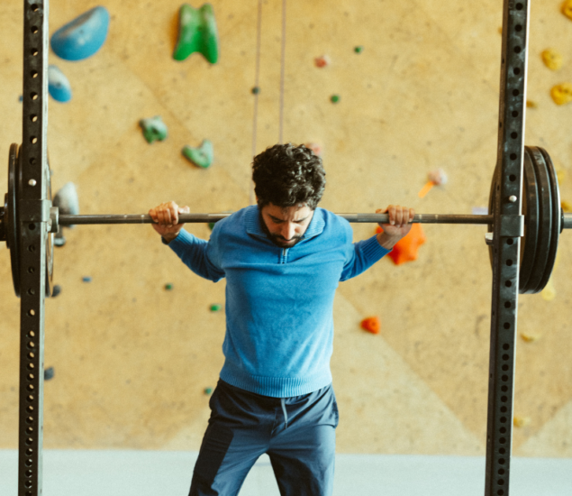 A man in a blue sweater lifts a loaded barbell on his shoulders in a gym with a climbing wall in the background.