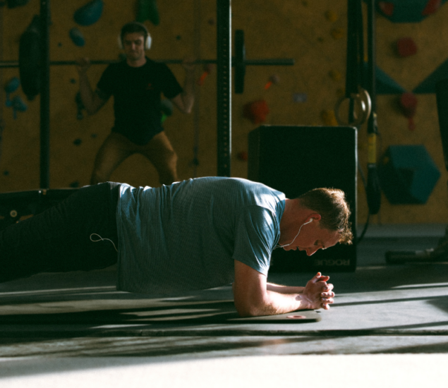 A man wearing headphones does a plank exercise on a gym mat while another person lifts weights in the background. The gym is dimly lit with climbing holds and gym equipment visible.