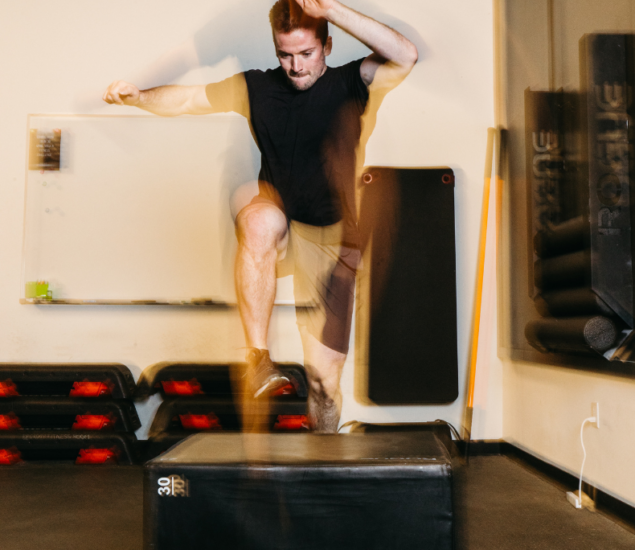 A man in athletic wear performs a high jump onto a black plyometric box in a gym. His movement appears as a blur, emphasizing speed and motion. Gym mats and equipment are visible in the background.