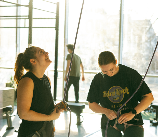 Two women in harnesses prepare for indoor climbing, smiling and standing near ropes in a sunlit gym with large windows. A person stands in the background by the window.