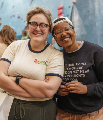 Two people stand close together indoors, smiling at the camera. One wears glasses and a cream-colored T-shirt, while the other wears glasses, a dark sweatshirt with text, and a cap. Blurry people and climbing walls are in the background.