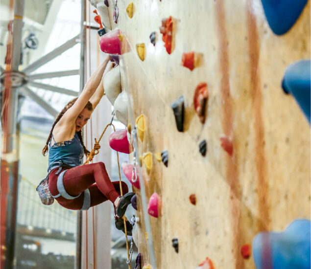 A woman with braided hair climbs an indoor rock wall, gripping colorful holds and wearing athletic gear with a harness and safety rope attached.