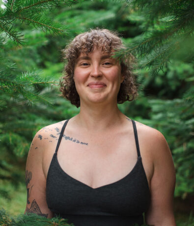A person with curly short hair and tattoos on their shoulder and arm stands outdoors among green pine branches, smiling and wearing a dark camisole top.