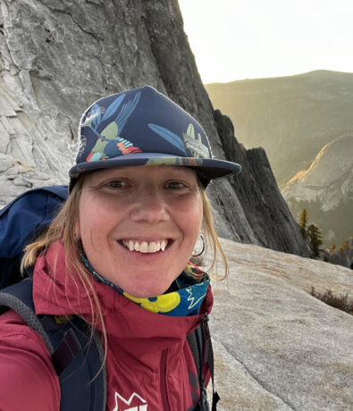 A smiling person wearing a cap, scarf, and backpack stands on a rocky mountainside with steep cliffs and a scenic valley in the background at sunset.