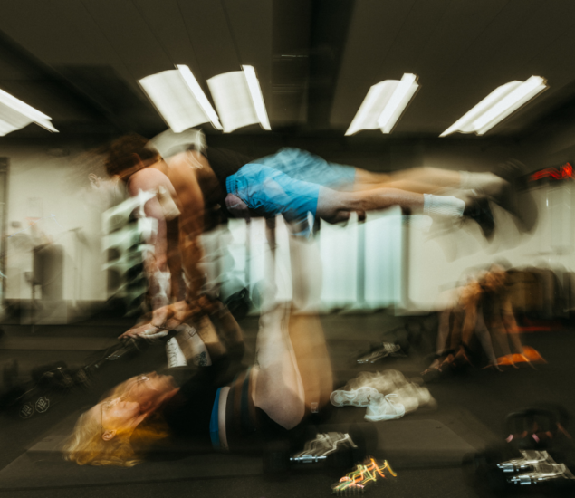 A blurry image of two people exercising indoors, with one person lying on their back and lifting another, creating a dynamic, climbing-inspired motion effect in a gym setting.