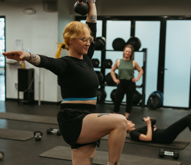 A woman in athletic wear lifts a kettlebell overhead with one arm while balancing on one leg in a gym, showcasing strength and balance essential for climbing. Two other women are in the background, one standing and smiling, the other lying on the floor.