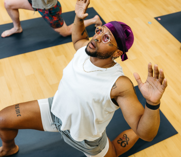 A person wearing glasses, a purple headscarf, and white workout clothes is in a low lunge yoga pose on a mat indoors, arms raised as if climbing towards new heights, looking upwards. Another person is partially visible behind them.