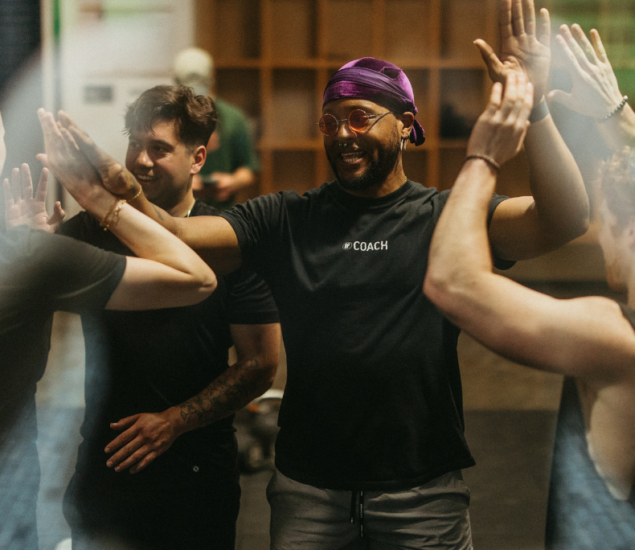 A group of people smiling and giving each other high fives in a climbing gym. One person in the center wears a purple headscarf, glasses, and a shirt that says COACH. They appear happy and energized.