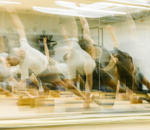 A group of people performing side plank yoga poses in a studio with mirrored walls; the image has a motion blur effect, creating overlapping shapes and dynamic movement, reminiscent of climbing fluidly through space.