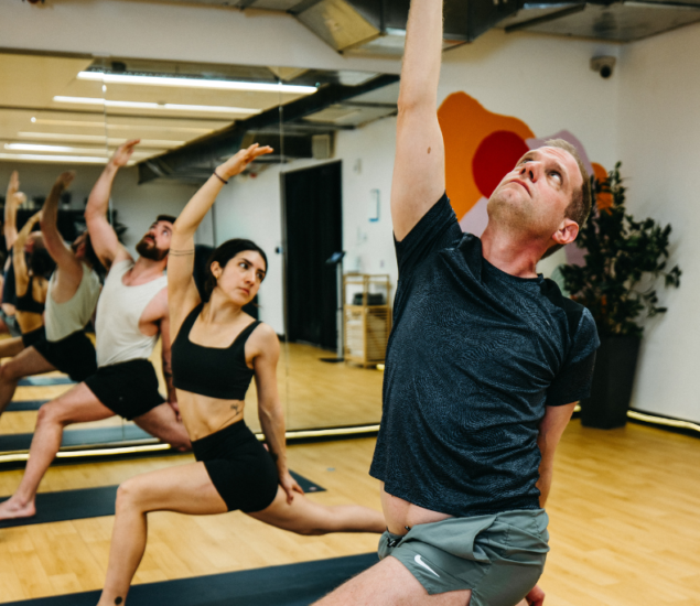 A group of people practice yoga in a studio, performing a lunge pose with one arm extended upward, as if reaching toward a climbing hold. The room features wooden floors, mirrors, and plants in the background.