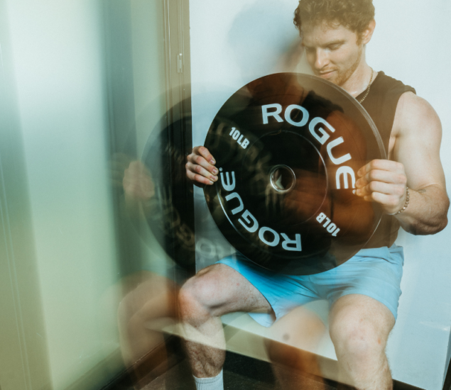 A man in a black tank top and blue shorts does a wall sit while holding a large ROGUE 10 lb weight plate vertically, as if preparing for climbing training. The motion blur effect makes his arms and the weight appear doubled.