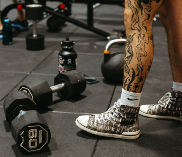 A person with tattooed legs and patterned sneakers stands near a 60-pound dumbbell, a kettlebell, and a water bottle in a gym setting, ready for their next climbing-inspired workout.