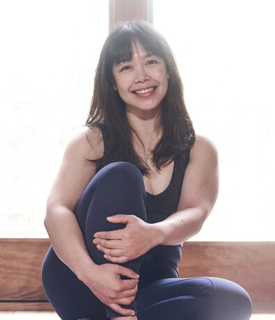 A woman with long dark hair, wearing a dark sleeveless top and leggings, sits cross-legged indoors, smiling warmly at the camera. Soft natural light comes through a window behind her.