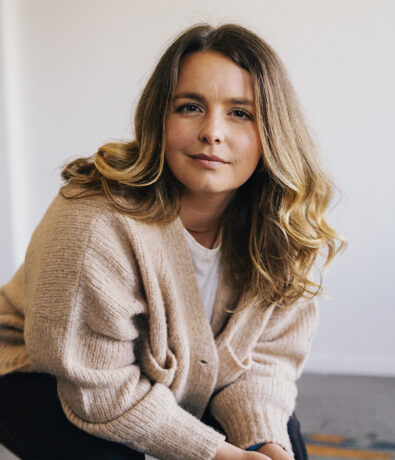 A woman with wavy, shoulder-length blonde hair wearing a beige cardigan and white shirt sits and looks at the camera, smiling softly, against a plain light background.