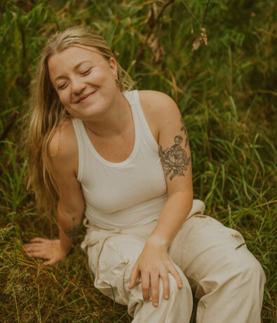 A smiling woman with long blonde hair sits on grass, wearing a white tank top and light pants. She has a floral tattoo on her upper arm and appears relaxed and content outdoors.