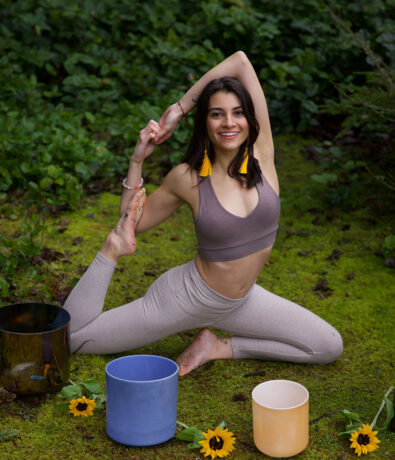 A woman practices yoga outdoors on green moss, smiling in a seated pose with one arm overhead and her foot in hand. She is surrounded by sunflowers and colorful singing bowls.