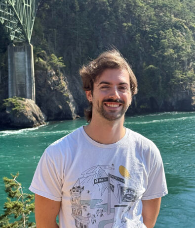 A smiling person with brown hair and a mustache stands in front of a turquoise river with a forested cliff and a large bridge structure in the background. They are wearing a white graphic T-shirt.