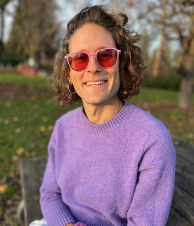 A person with wavy brown hair wearing pink sunglasses and a purple sweater smiles while sitting on a wooden bench in a park with autumn trees and scattered leaves in the background.