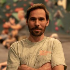 A man with short brown hair and a beard stands with his arms crossed and smiles slightly. He is wearing a light-colored t-shirt at an indoor climbing gym with blurred people and climbing walls in the background.