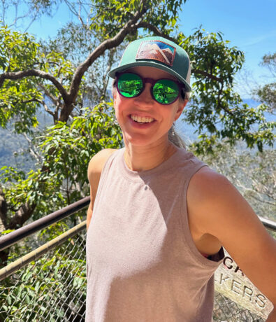 A smiling person in sunglasses and a cap stands outdoors by a railing, surrounded by green trees and sunlight, with mountains visible in the background.