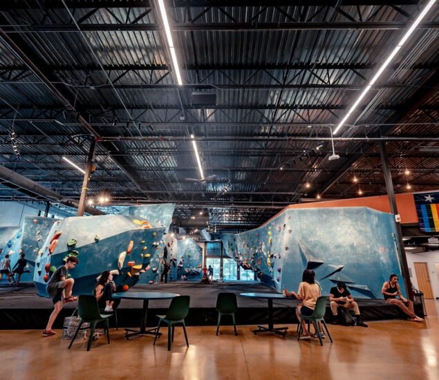 Indoor rock climbing gym with several people climbing on blue bouldering walls and others sitting at green chairs and tables. Overhead lights illuminate the Austin Climbing Gym, and a pride flag is visible on the wall to the right.