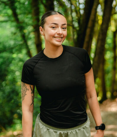 A woman with dark hair in a bun, wearing a black athletic shirt and light shorts, smiles outdoors on a forest trail. She has a tattoo on her left arm and is wearing a smartwatch. Green trees are in the background.