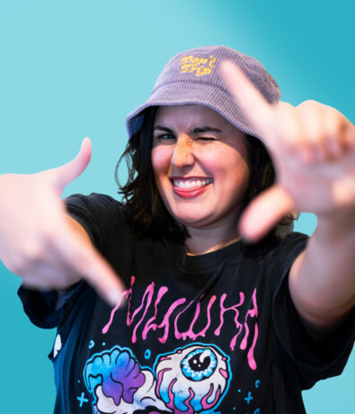 A smiling person in a purple bucket hat and a graphic T-shirt winks and forms a rectangle with their fingers, posing playfully in front of a turquoise background.