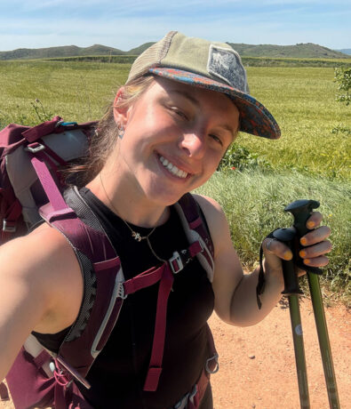 A smiling person wearing a cap and backpack takes a selfie while hiking on a dirt trail, holding trekking poles. Green fields and hills are visible in the background under a sunny sky.