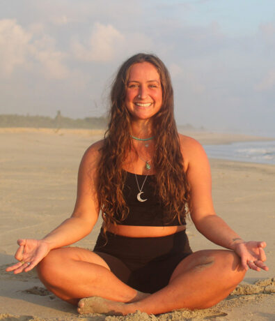 A woman with long brown hair sits cross-legged on a sandy beach, smiling and holding a relaxed, open-handed meditation pose. She wears a black outfit and layered necklaces. The sky is partly cloudy.