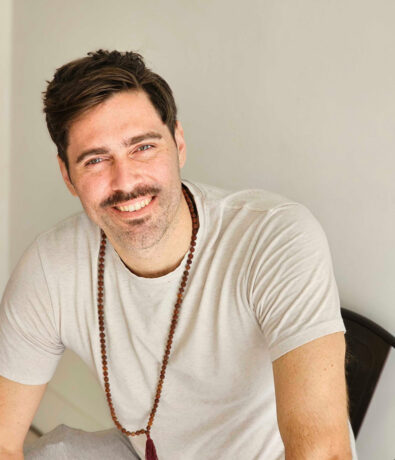 A man with short brown hair and a mustache, wearing a light gray t-shirt and a beaded necklace, sits and smiles at the camera against a plain light-colored background.