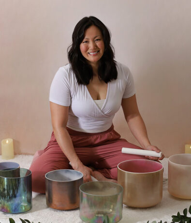 A smiling woman sits cross-legged on a mat, surrounded by colorful crystal singing bowls and candles. She holds a mallet, ready to play the bowls, suggesting a sound healing or meditation session.