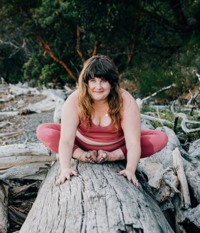 A woman wearing a pink sports outfit sits on a large log outdoors, doing a yoga pose with her legs crossed and hands on the log, smiling at the camera. There are trees and driftwood in the background.