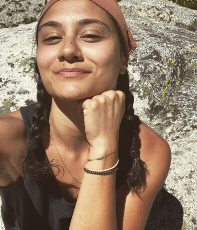 A person with two braided pigtails, wearing a brown headband and a sleeveless black top, rests their chin on their fist and smiles slightly while sitting outdoors on a sunlit rock.