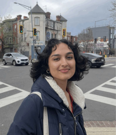 A person with short, wavy dark hair and a blue jacket stands at a city street intersection near the DC Rock Climbing Gym, smiling at the camera. Buildings, cars, and traffic lights are visible in the background under a cloudy sky.