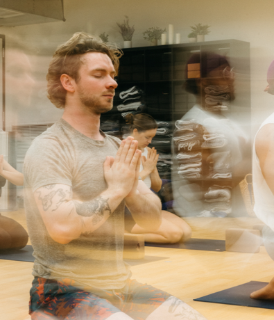 A group of people practice yoga indoors, kneeling on mats with hands pressed together in prayer position. The image has a motion blur effect, creating a sense of calm movement and focus.