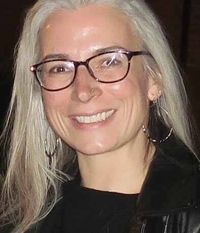 A smiling woman with long gray hair and glasses wears a black top and leather jacket, standing indoors at the DC Rock Climbing Gym against a dark background.