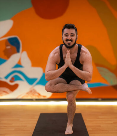 A man in athletic wear balances on one leg in a yoga pose at DC Rock Climbing Gym, smiling with hands in prayer position. A colorful mural with abstract shapes and figures is in the background.