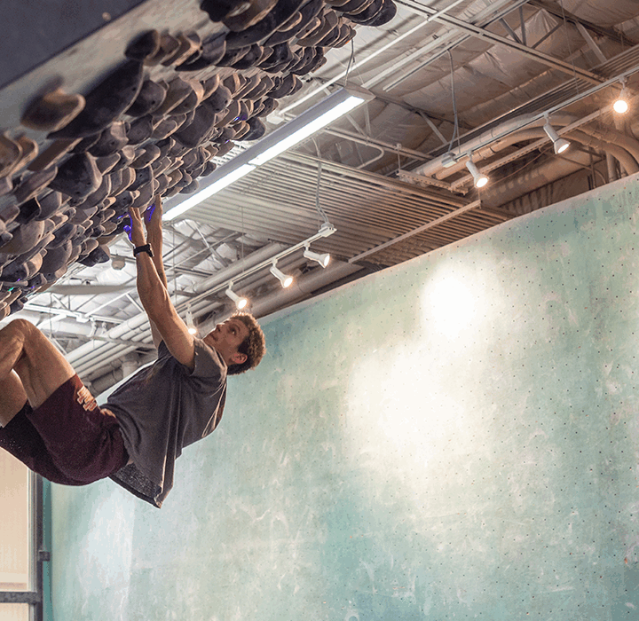 A person climbs an overhanging indoor bouldering wall at an Austin climbing gym, gripping holds with both hands and feet. The gym features high ceilings, overhead lights, and a green climbing wall in the background.