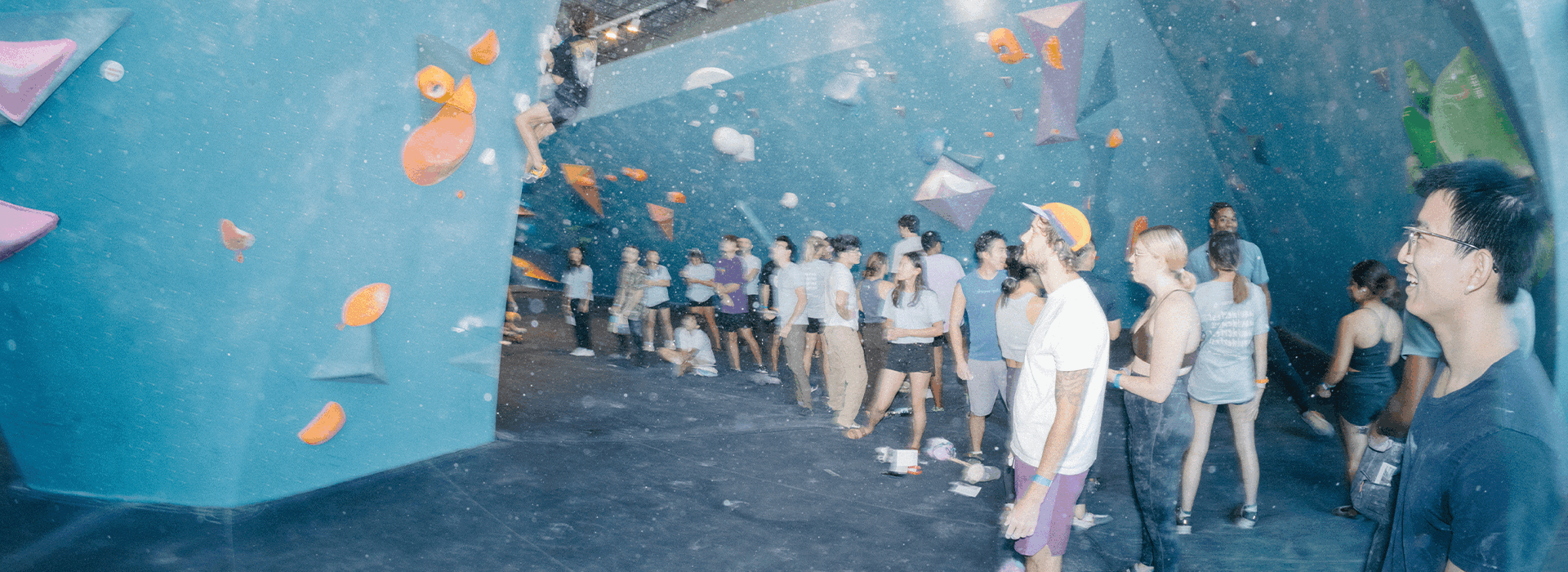 A group of people stand and watch climbers at an Austin climbing gym with blue bouldering walls and colorful holds. Some climb, while others observe and chat, creating a lively and social atmosphere.