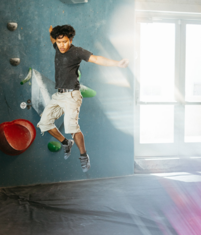 A person in climbing shoes and shorts jumps off the ground at an indoor climbing gym, surrounded by colorful climbing holds and a padded mat. Sunlight streams through a door in the background, capturing an energetic moment at Brooklyn Climbing Yoga and Fitness.