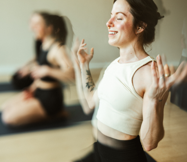A woman sits cross-legged on a yoga mat at Brooklyn Climbing Yoga and Fitness, smiling with her eyes closed and hands raised. Another person is visible in the blurred background, both in a peaceful indoor setting.