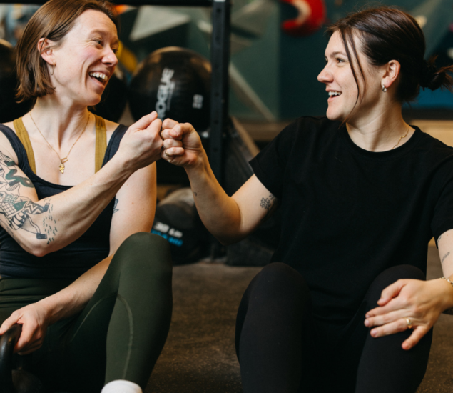 Two women sit on the Brooklyn Climbing Yoga and Fitness gym floor, smiling and giving each other a fist bump. Both wear athletic clothing, with gym equipment in the background. They appear happy and relaxed.