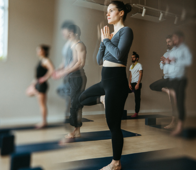 A group of people practice yoga indoors at Brooklyn Climbing Yoga and Fitness, standing on mats in tree pose with hands together at their chests. The focus is on a woman in the foreground, with others slightly blurred in the background.