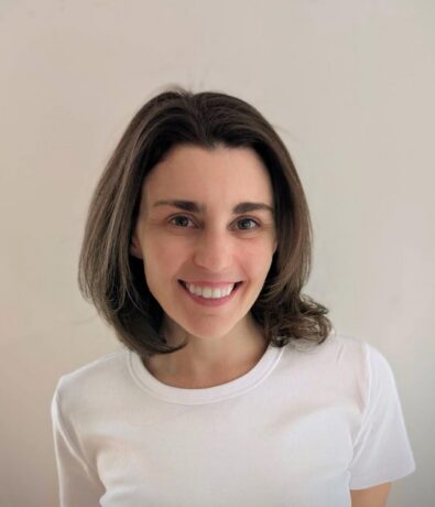 A woman with shoulder-length brown hair and a white t-shirt smiles at the camera against a plain, light-colored background, embodying the vibrant spirit of Brooklyn Climbing Yoga and Fitness.