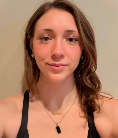 A woman with wavy brown hair wearing a black top and two gold necklaces, one with a rectangular pendant, smiles softly at the camera against a plain light background, radiating the calm confidence found at Brooklyn Climbing Yoga and Fitness.