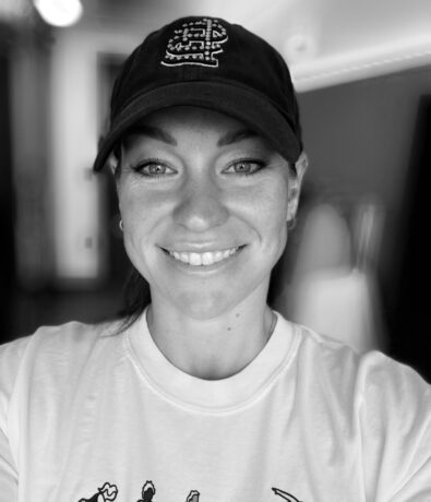A woman smiles at the camera, wearing a dark baseball cap and a light-colored T-shirt with yoga poses illustrated on it. The photo is in black and white, with a blurred background.