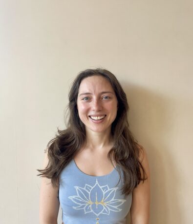 A woman with long brown hair wearing a blue crop top with a lotus flower design stands smiling against a plain beige wall, embodying the vibrant spirit of Brooklyn Climbing Yoga and Fitness.